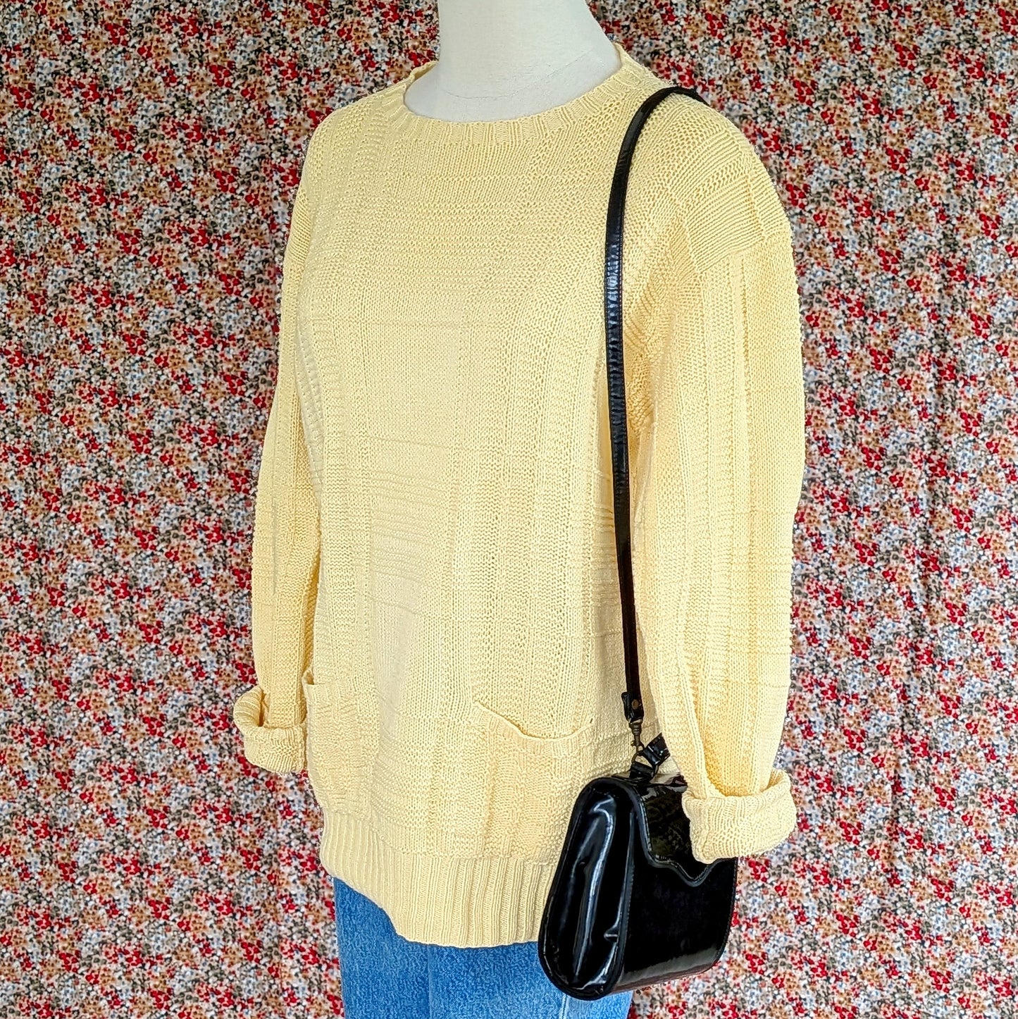 Mannequin wearing a yellow sweater, blue jeans, and a black handbag against a floral patterned wall.